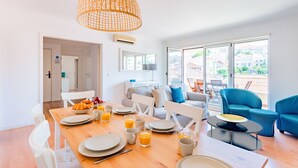 Une salle à manger lumineuse avec une table en bois dressée pour le petit-déjeuner, une grande fenêtre donnant sur des immeubles et une chaise bleue à motifs.
