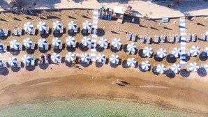 Vista aérea de una playa con hileras de sombrillas y sillas de playa, una torre de salvavidas y aguas poco profundas y cristalinas.
