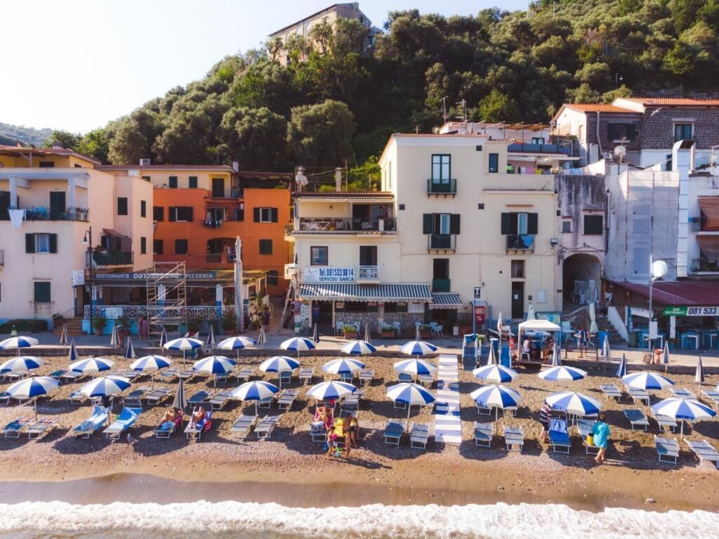 Una playa con hileras de sombrillas y sillas de descanso, un restaurante y edificios con balcones.