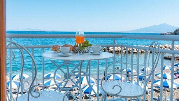 Un balcón con una mesa dispuesta con comida y bebidas, con vistas a una playa con sombrillas y sillas.