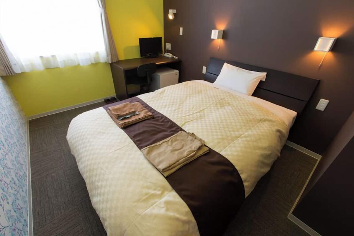 A hotel room with a bed, desk, and chair, a small desk lamp, and a wall-mounted light fixture.