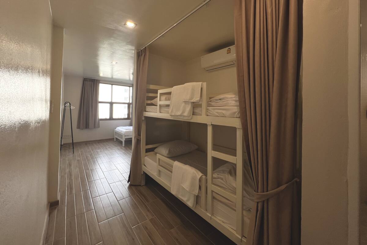A dormitory room with bunk beds, a window with curtains, a radiator, and a small table.
