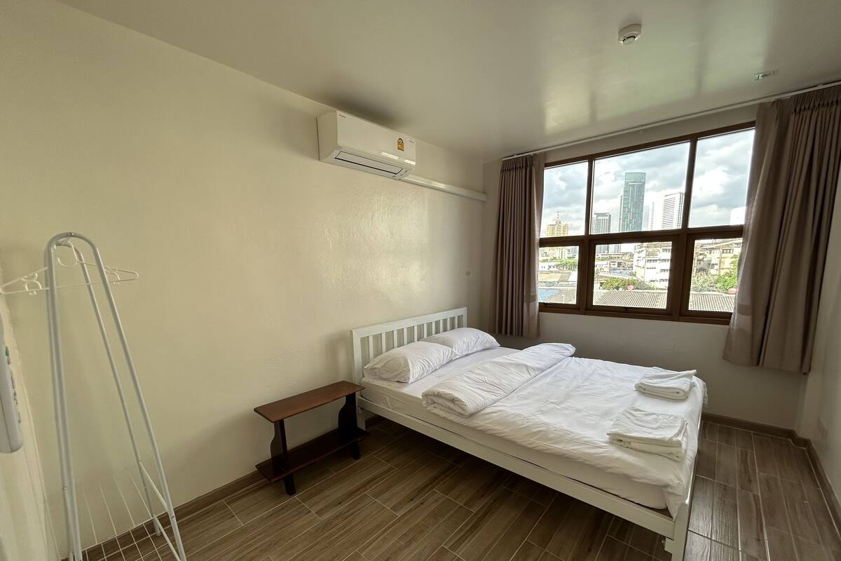 A bedroom with a bed, a small bedside table, a window with curtains, and an air conditioning unit on the wall.