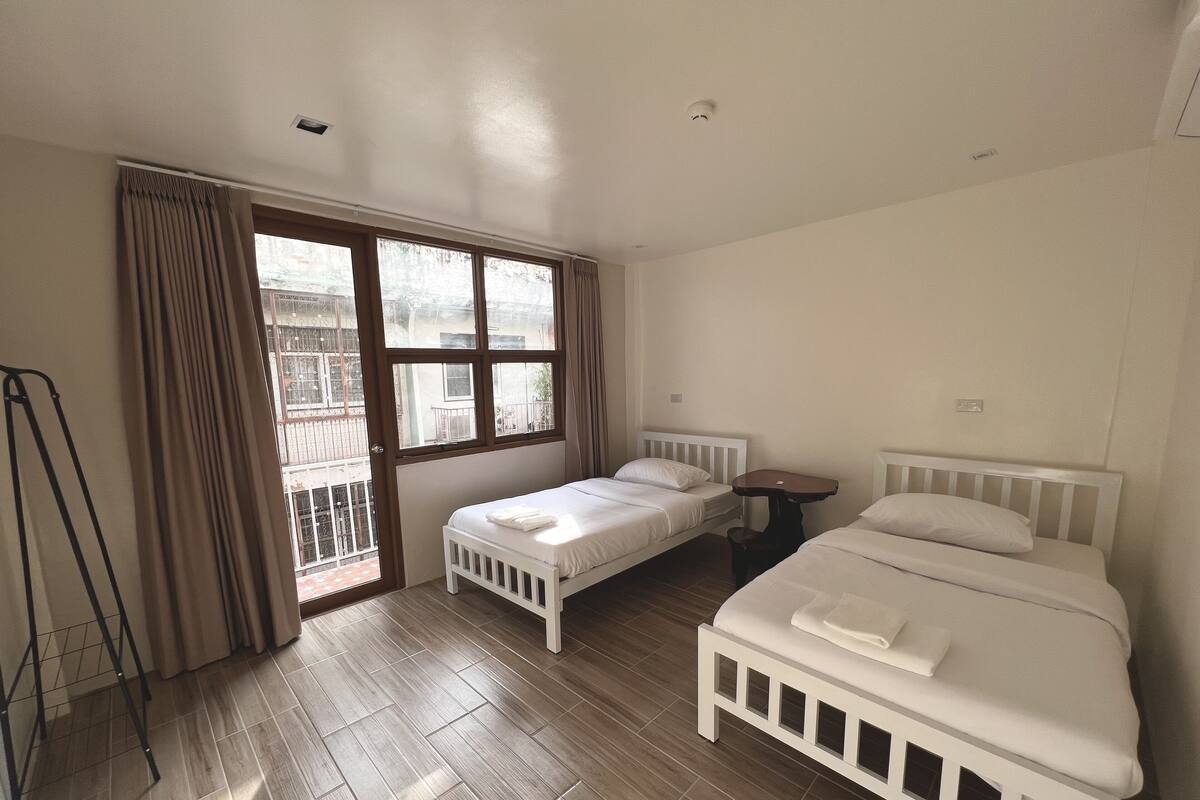 A bedroom with two single beds, a small table, and a window with curtains.