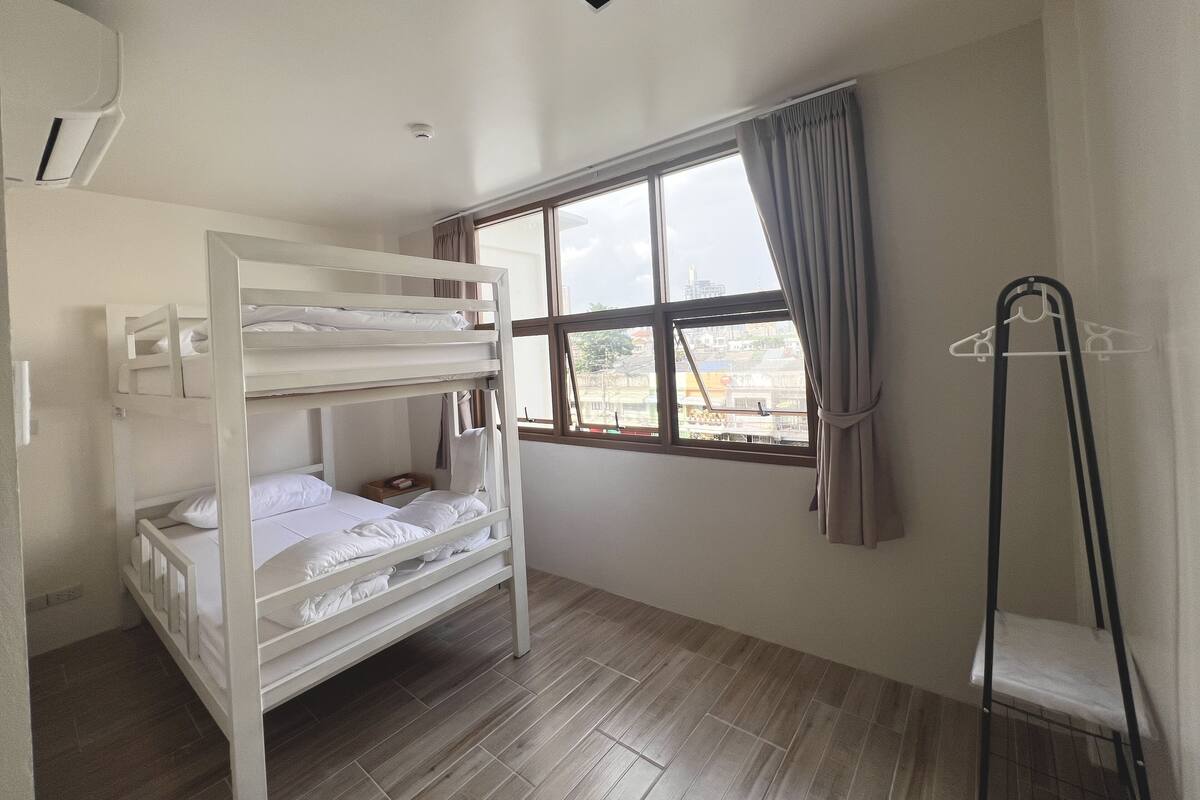 A room with a bunk bed, a window with curtains, a hanging clothes rack, and a ceiling vent.