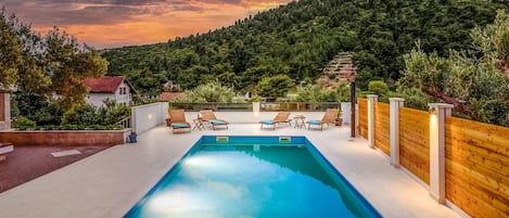 A rectangular swimming pool with lounge chairs and a wooden fence, surrounded by lush greenery and a mountainous backdrop.