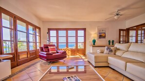 A spacious living room with a red armchair, beige sofa, and a coffee table with magazines. Large windows offer a view of the ocean.