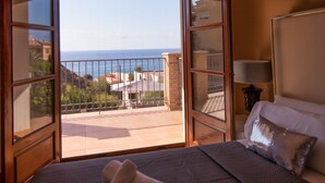A hotel room with a balcony overlooking the sea, a bed with a pillow, and a nightstand with a lamp.