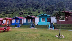 A row of colorful cabins with a grassy area in front and a forested hill in the background. - PRIDE INN LUXURY RESORT BY UK HEAVEN (Yamkeshwar)