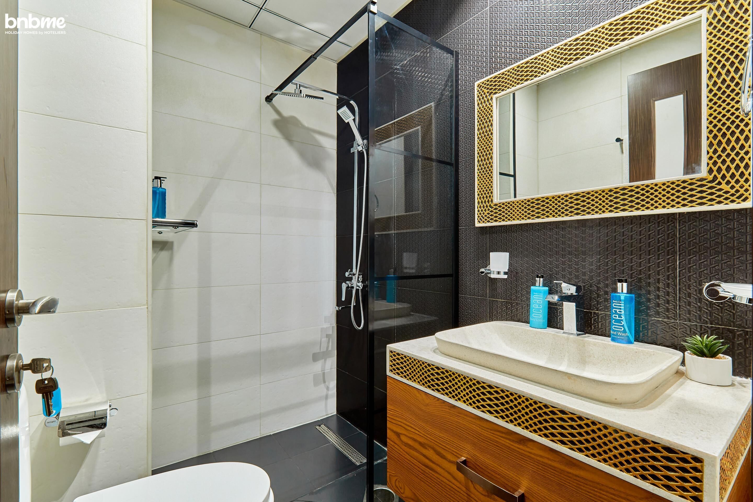 Un baño moderno con ducha envuelta en vidrio, tocador de madera con lavabo y un espejo grande con marco decorativo.