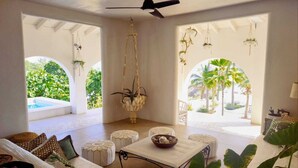 Un amplio espacio de sala con ventilador de techo, un conjunto de sofás, otomanas y plantas colgantes.