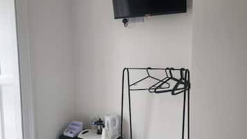Habitación de hotel con una cama, un aparador negro con teléfono y hervidor, un televisor montado en la pared y un tendedero de metal con toallas.