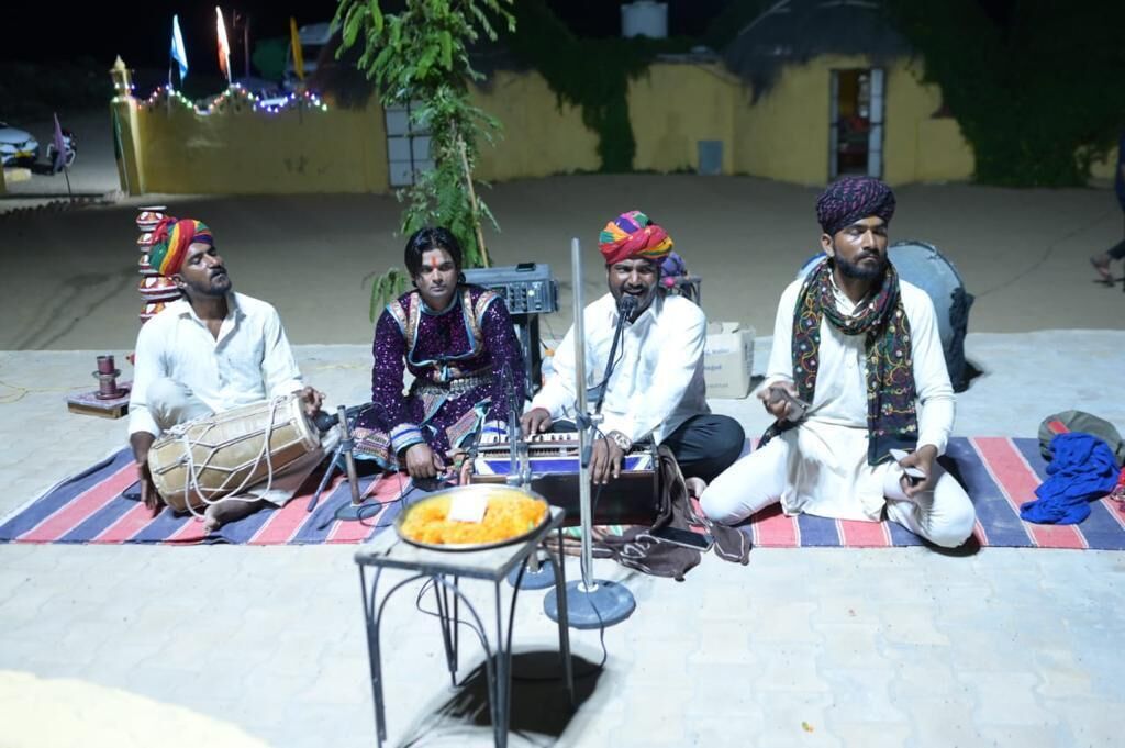 Cuatro personas sentadas sobre una estera con instrumentos musicales, sugiriendo un evento cultural o musical.