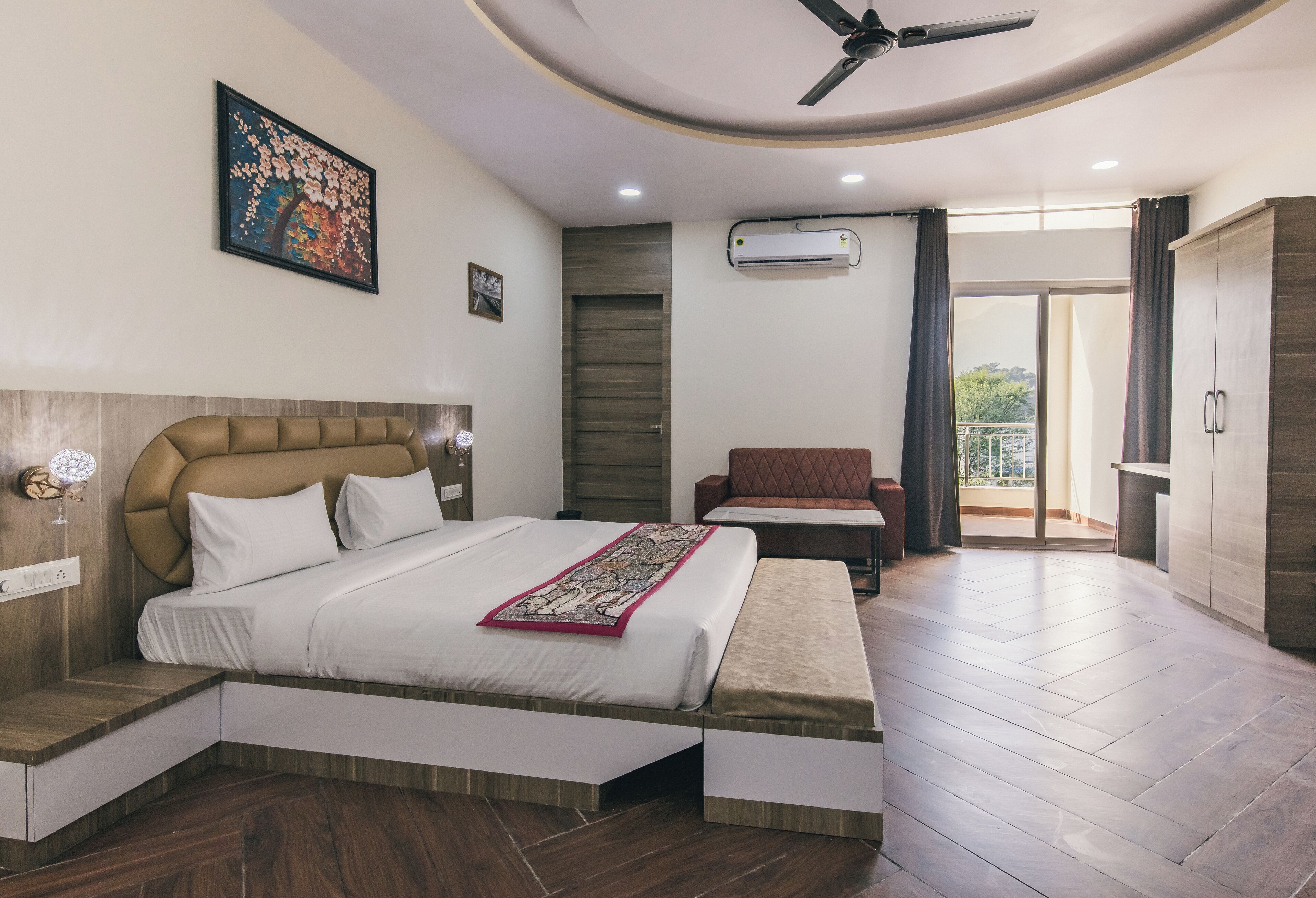 Une chambre moderne avec un grand lit, un ventilateur de plafond, un appareil de climatisation, une petite table et une vue sur l’extérieur.