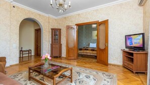 Un salon avec une table basse en bois, un téléviseur et un canapé. On y trouve également un lustre, un meuble en bois et une porte qui donne sur une autre pièce.