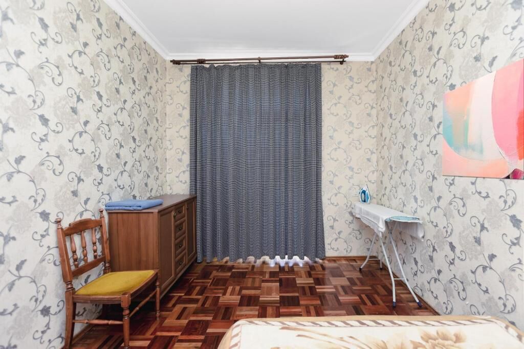 A room with patterned wallpaper, a wooden chair with a yellow cushion, a bed with a floral bedspread, and a radiator.