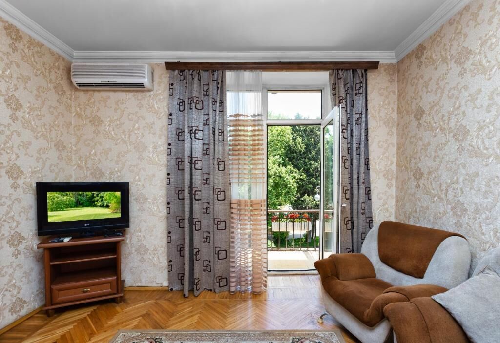 Un salon avec un téléviseur posé sur un meuble en bois, un canapé marron, des rideaux à motifs et une porte-fenêtre coulissante donnant sur un balcon.