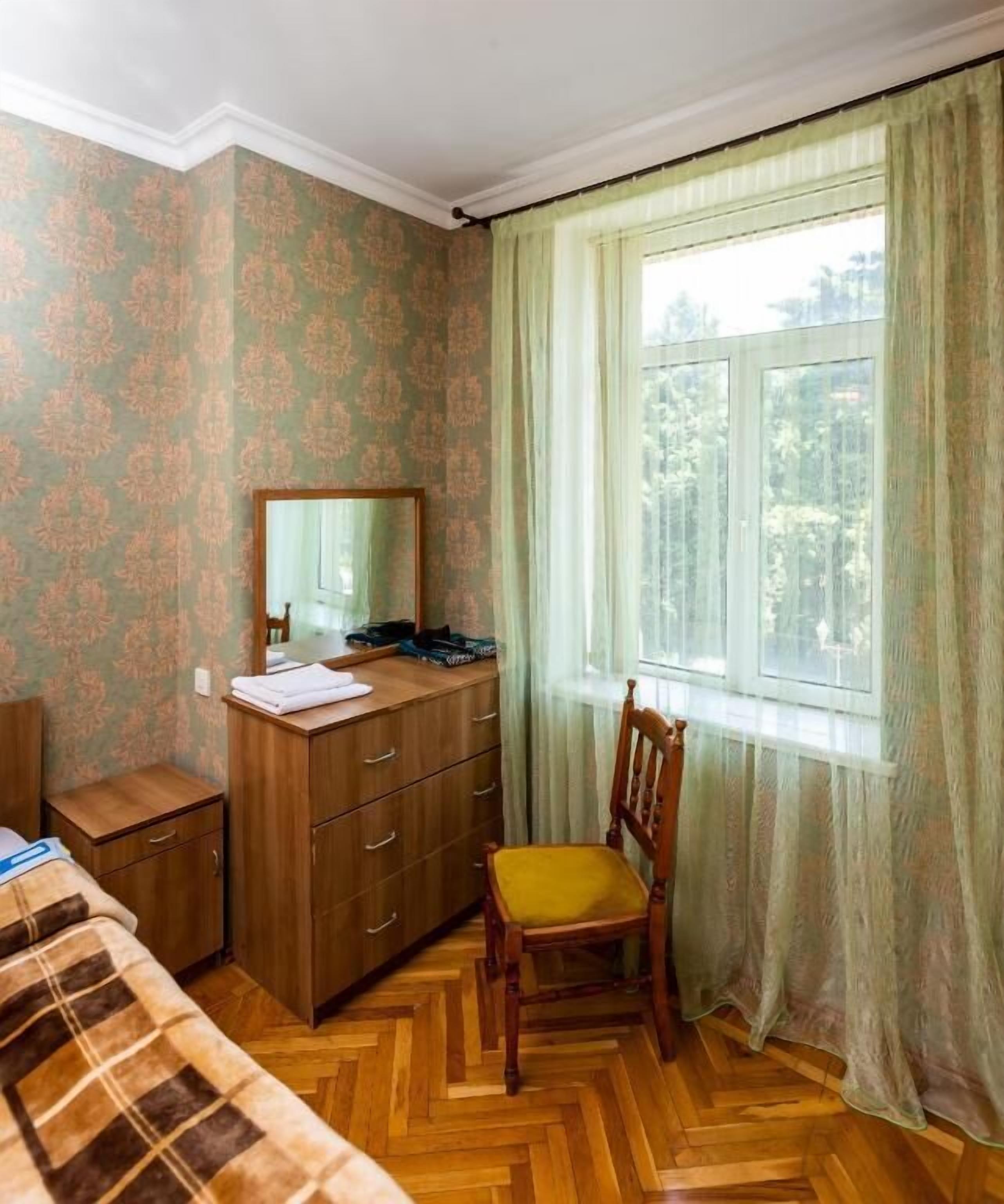 Een kamer met een houten dressoir, een stoel, een bed met een geruit plaid en een raam met groene gordijnen.