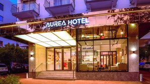 Entrada de hotel con fachada de vidrio, mobiliario exterior y una recepción visible.