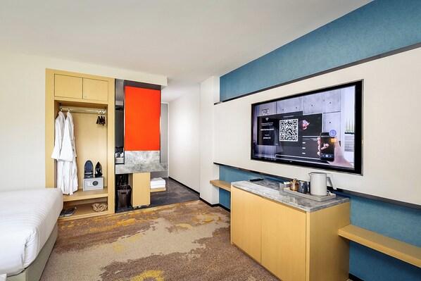 Una habitación de hotel moderna con una cama, un armario, una kitchenette pequeña y un televisor en la pared que muestra un código QR.