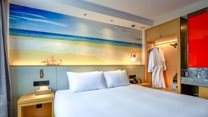 Una habitación de hotel con una cama grande, una mesita de noche con una lámpara, una bata colgada en un perchero y un mural en la pared con un paisaje de playa.