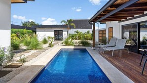 Una piscina exterior moderna con cubierta de madera, rodeada de vegetación y un cielo despejado.