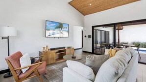 Amplia sala de estar con televisor grande, un sofá, una silla y comedor con vistas al exterior.