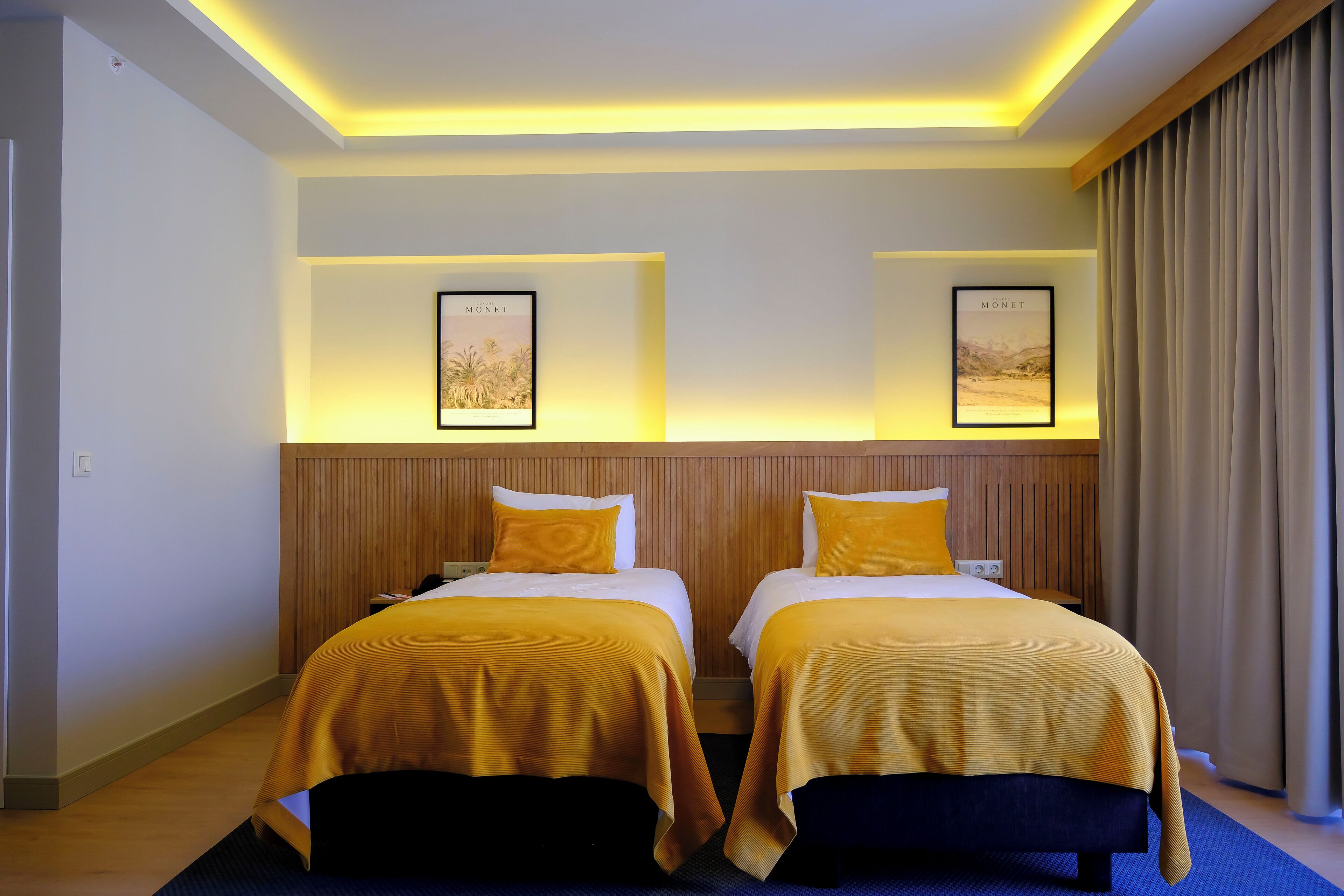 Ein Hotelzimmer mit zwei Betten, jeweils mit gelber Bettdecke, einem Holz-Kopfteil und zwei gerahmten Bildern an der Wand.