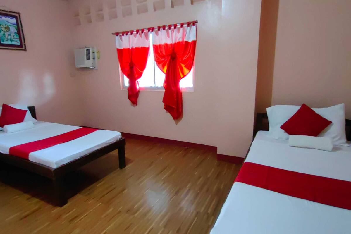 Ein Zimmer mit zwei Betten, jeweils mit weißer Bettwäsche und roten Akzenten, einem Holzboden, einem Fenster mit roten Vorhängen und einer wandmontierten Klimaanlage.