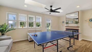 A room with a ping pong table, ceiling fan, windows, and a fireplace.