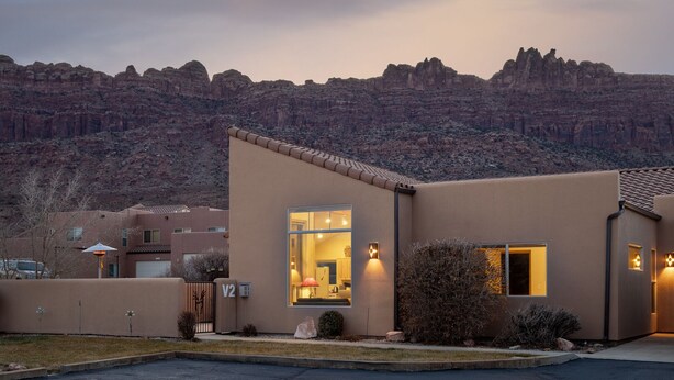 Una casa moderna de dos pisos con un ventanal grande y un interior bien iluminado, rodeada de un paisaje desértico.