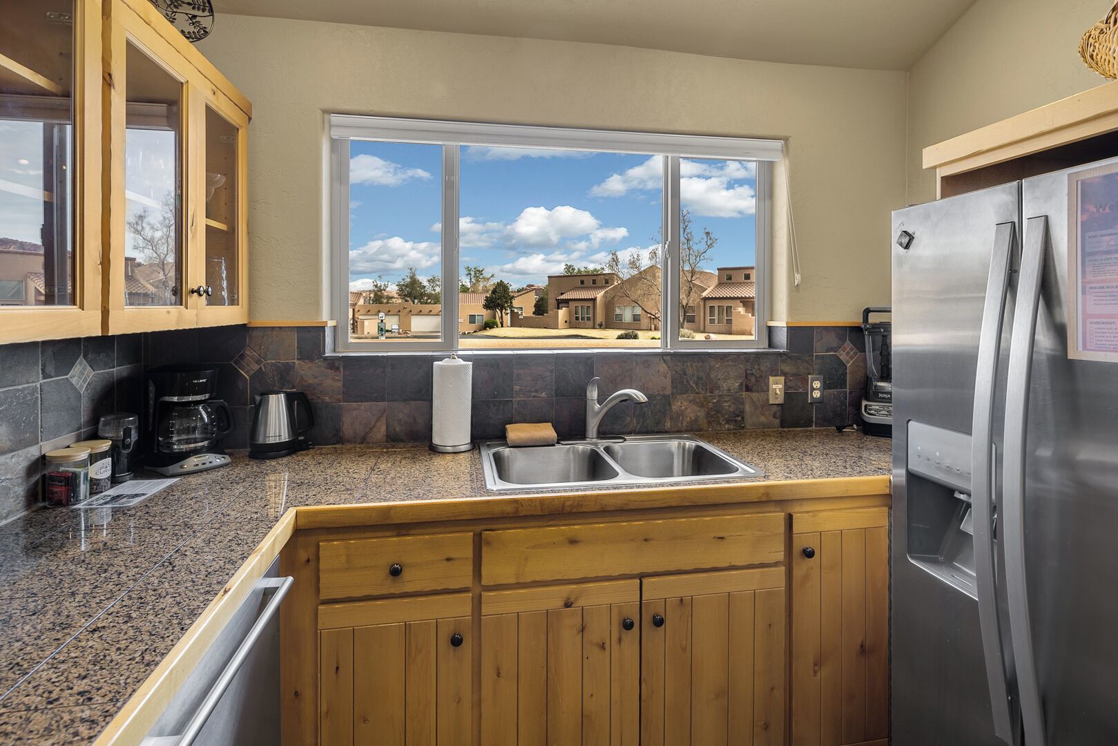 A kitchen with granite countertops, wooden cabinets, a stainless steel refrigerator, and a window offering a view of houses.