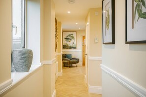 A hallway with a patterned vase on a windowsill, a framed artwork on the wall, and a chair in the distance.