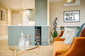 Una sala de estar moderna con una mesa de centro de vidrio, un sofá con cojines, una zona de cocina con un refrigerador y una mesa de comedor.