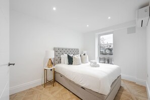A modern bedroom with a large bed, a bedside table with a lamp, and a view of the city through a window.