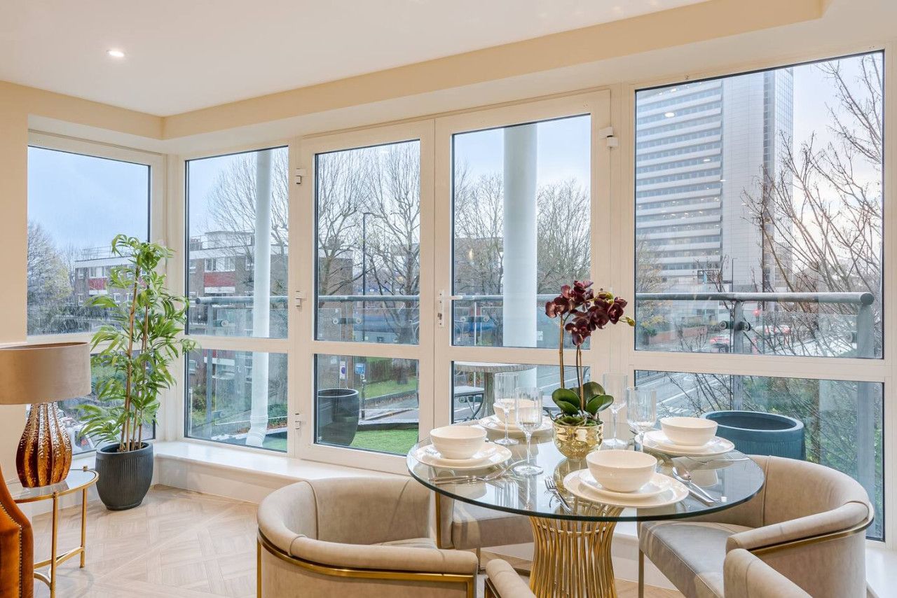 A modern dining area with a glass table set for four, surrounded by large windows offering a view of the cityscape.