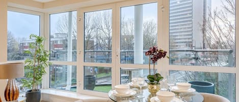 A modern dining area with a glass table set for four, surrounded by large windows offering a view of the cityscape.