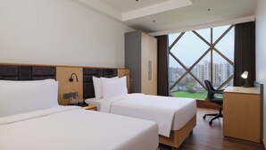 A hotel room with two beds, a desk, a chair, and a large window offering a view of buildings. - Regenta - The World Surat (Surat)