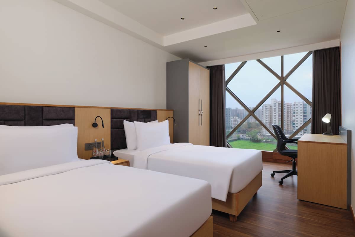 Habitación de hotel con dos camas, un escritorio, una silla y un ventanal con vistas a edificios.