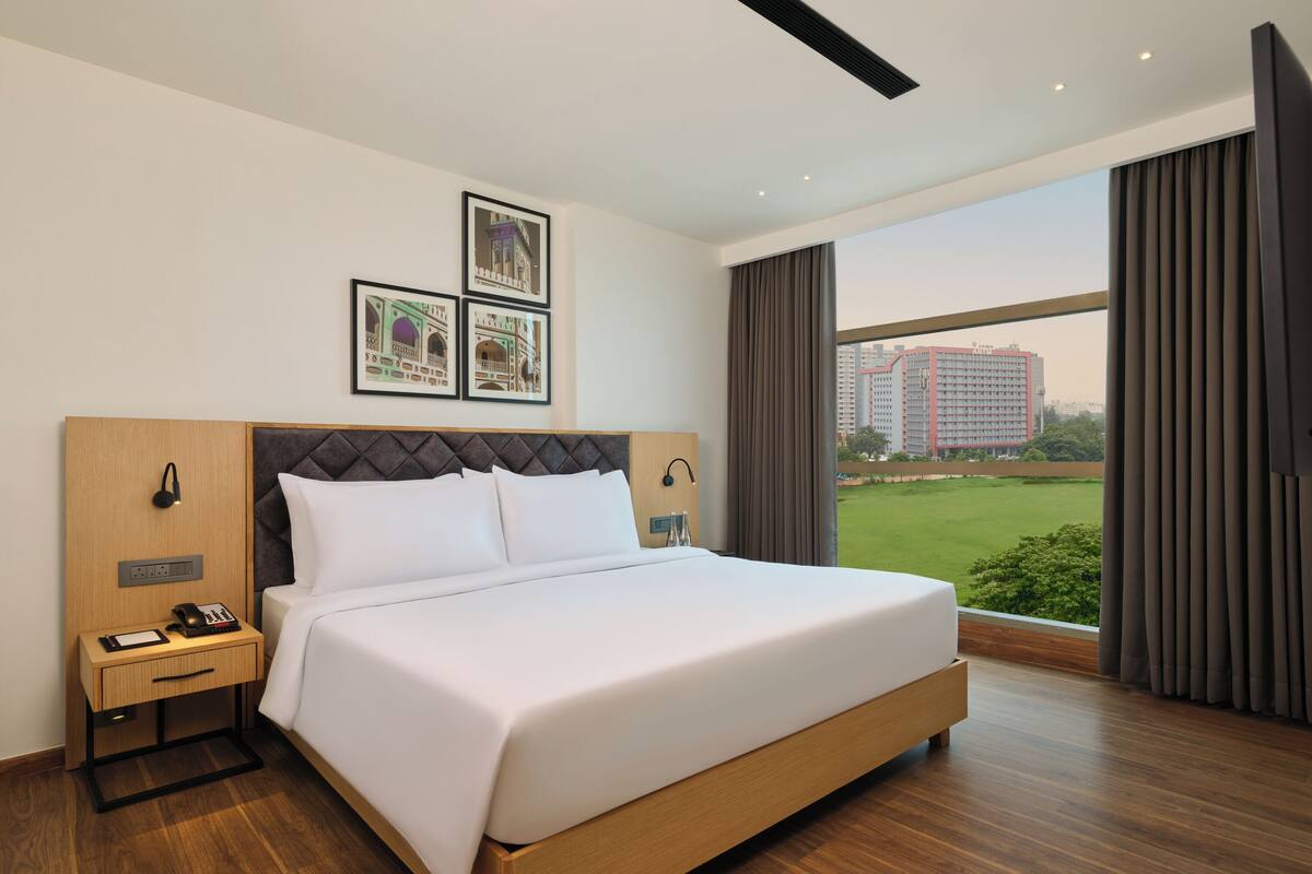 Habitación de hotel moderna con una cama grande, una mesita de noche con teléfono y cargador, una lámpara de noche y una ventana con vistas a edificios y vegetación.