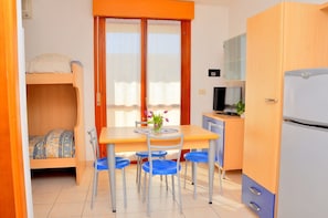 Une chambre dâhĂŽtel compacte comprenant un lit superposĂ©, une table Ă manger avec des chaises, un tĂ©lĂ©viseur, un rĂ©frigĂ©rateur et une fenĂȘtre avec des rideaux.