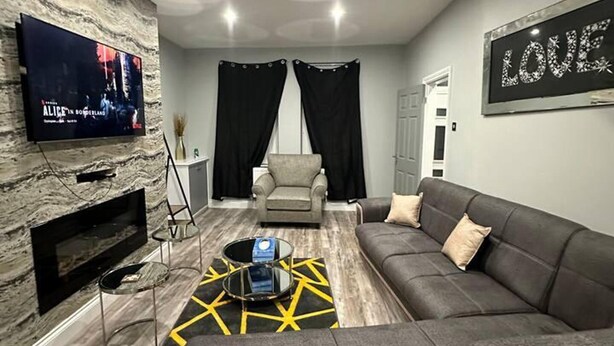 A modern living room with a grey sofa, a TV mounted on a textured wall, and a 'LOVE' sign on the wall. - Immaculate 4-bed Detached House in Bootle (Bootle)