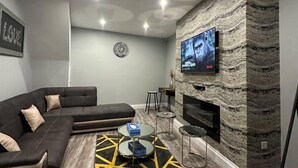 A modern living room with a sectional sofa, a flat-screen TV mounted on a stone wall, a coffee table with a geometric rug, and a small side table. - Immaculate 4-bed Detached House in Bootle (Bootle)