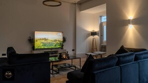 Ein modernes Wohnzimmer mit einem großen Flachbildfernseher, dunkelblauen Sofas, einem Couchtisch und einer Stehlampe.