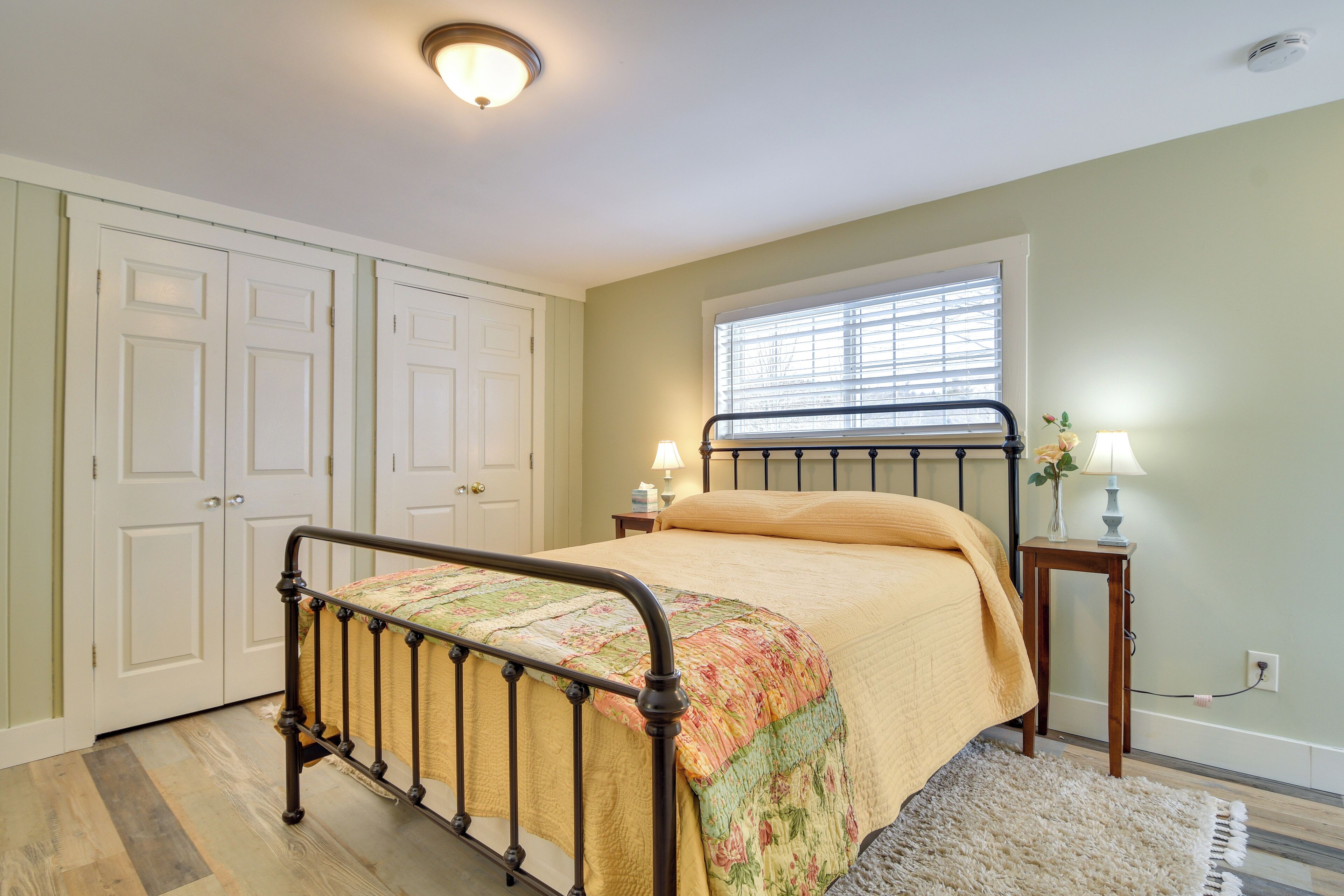 A bedroom with a bed, two bedside tables, a window with blinds, a door, and a ceiling light.