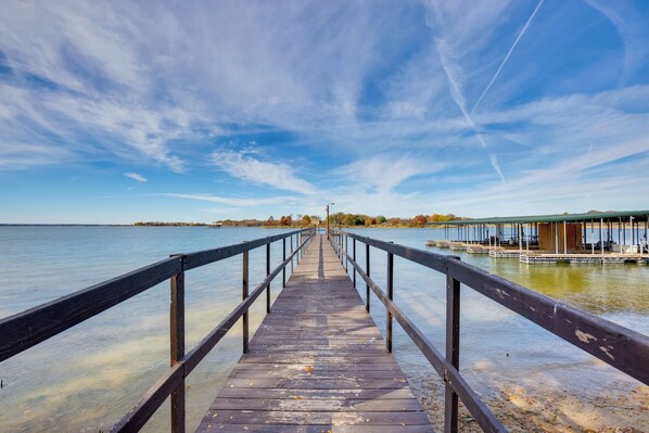 A wooden pier extending into calm water, with a clear sky above and a distant shoreline with trees and a building.