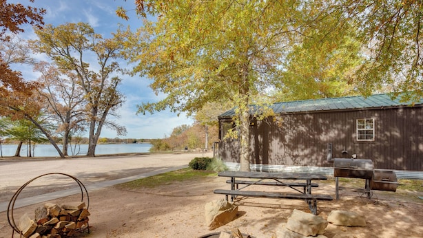 A rustic cabin with a picnic table and fire pit, surrounded by trees and a lake view. - Reservoir & Dock Access: Pet-friendly Kerens Cabin (Kerens)