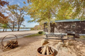 A rustic cabin with a picnic table and fire pit, surrounded by trees and a lake view.
