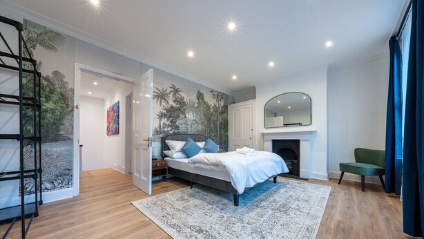 A modern bedroom with a bed, a fireplace, a green chair, and a large mirror. - West London Period Home (London)