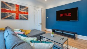 A modern living room with a blue accent wall, a large flat-screen TV, a grey sofa, and a patterned rug. - West London Period Home (London)